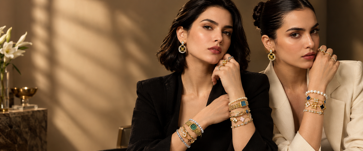 Two women in elegant blazers pose together, showcasing a variety of gold-toned jewelry including bracelets, rings, and earrings with colorful gemstone accents in a warm, sunlit interior setting.