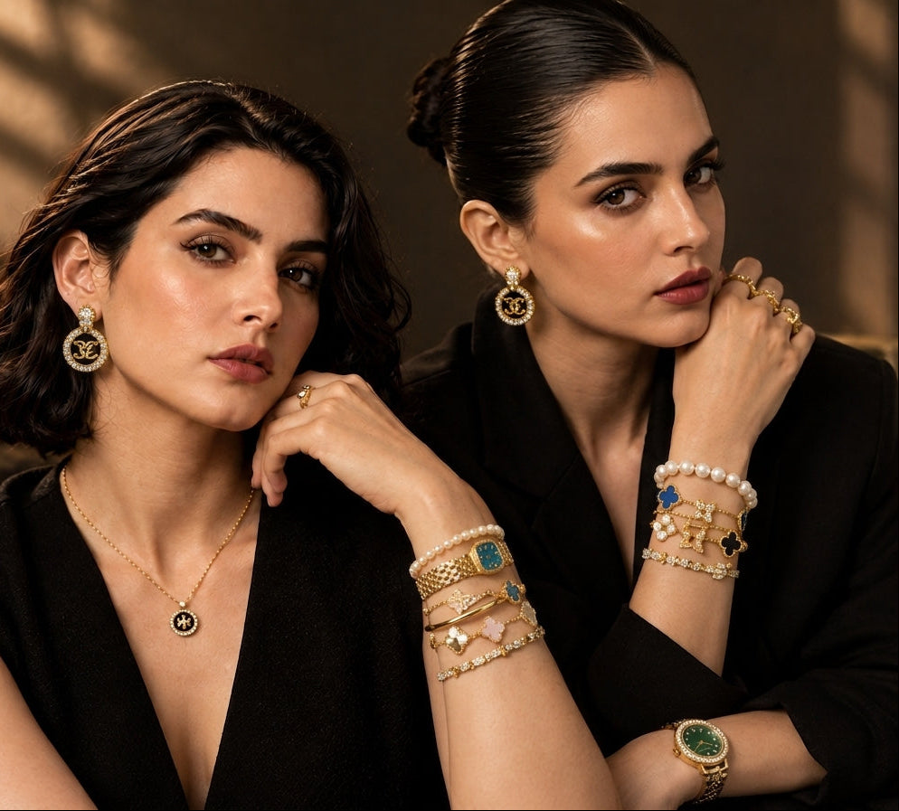 Two women in black outfits posing with luxury jewelry, including gold necklaces, bracelets, rings, and earrings, in a warm, moody indoor setting.
