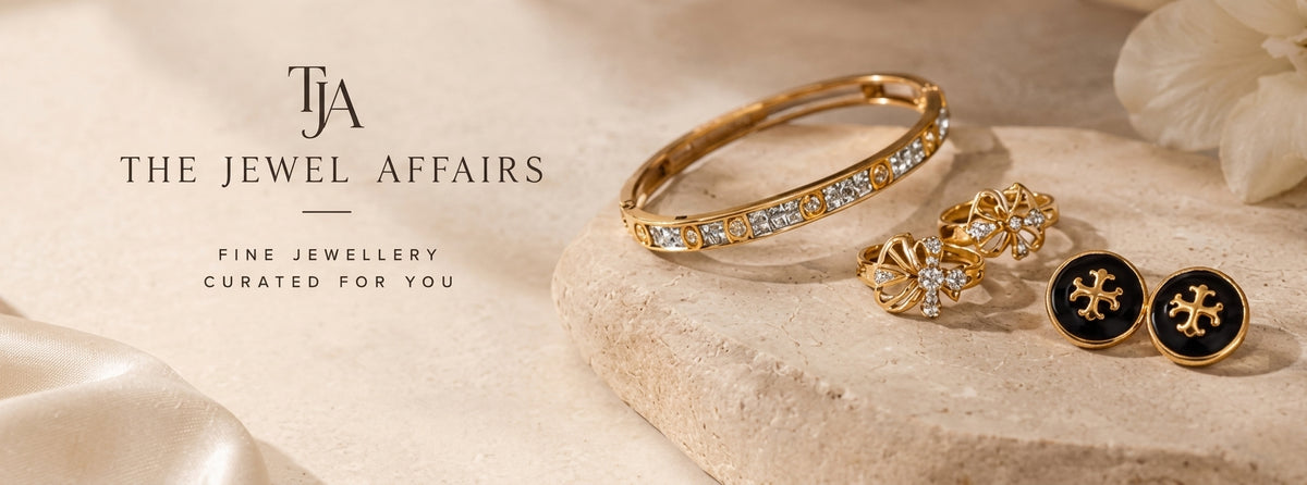 Gold jewelry set from The Jewel Affairs, including a bangle bracelet, a pair of floral stud earrings, and round black-and-gold button-style earrings, arranged on a stone slab with a soft beige background and brand text on the left.